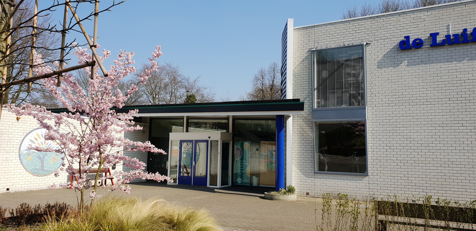 De ingang van de Luifel met het mozaïek, een struik in bloei met roze bloemen en de letters in blauw de Luifel op het gebouw