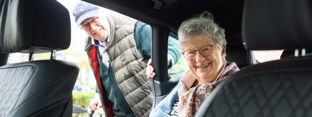Een lachende mevrouw zit in de auto van een vrijwilliger van vervoer door vrijwilligers, je ziet de binnenkant van de auto, met erachter de vrijwiliger.