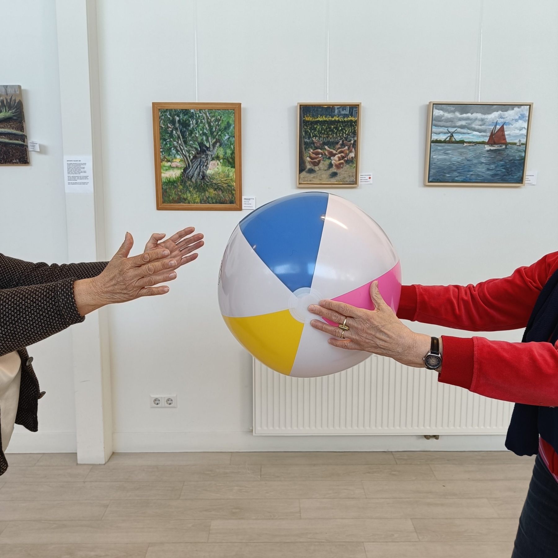 twee personen, waarvan je alleen de handen en armen ziet, die overgooien met een opblaasbare bal, met op de achtergrond een witte muur met schilderijen van een boom, kippen en een zeilboot met roodbruin zeil